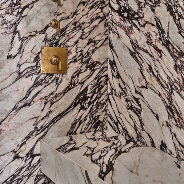Close-up of marble-lined shower with brass fixtures showing white marble stone with black and pink veining, and square brass shower control.