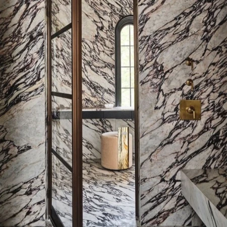 Luxurious bathroom interior featuring marble walls, a glass shower enclosure, a plush stool, and elegant brass fixtures, exuding sophisticated design.