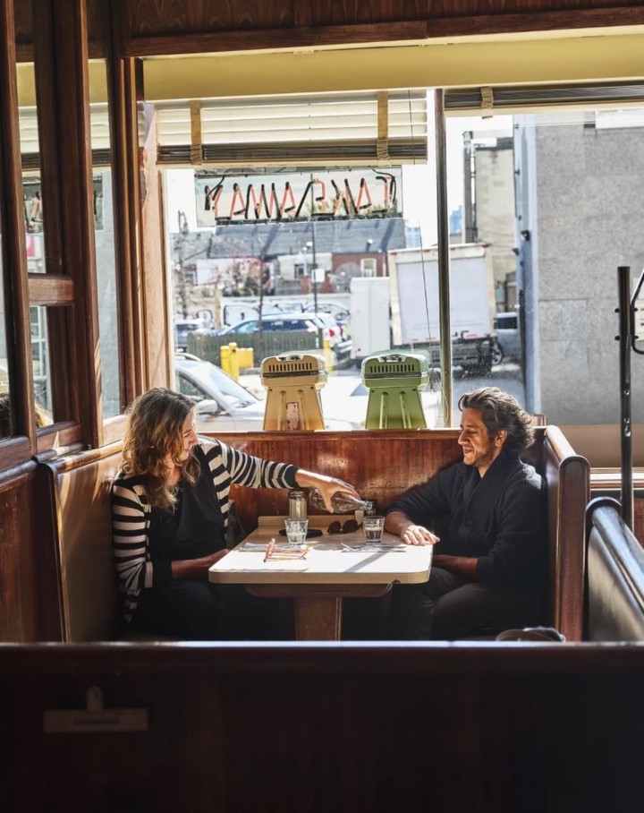 Two adult women sit at a wooden booth table inside a diner with beverages. Sunlight illuminates the urban setting and interior.