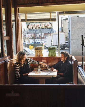 Two adult women sit at a wooden booth table inside a diner with beverages. Sunlight illuminates the urban setting and interior.