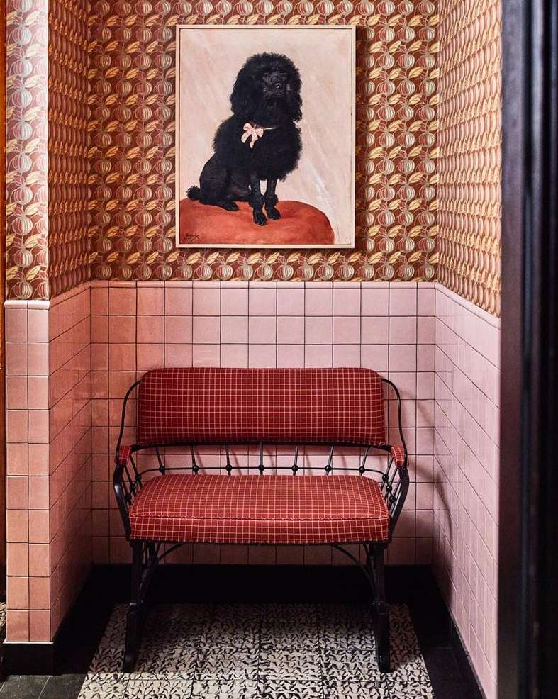Interior of a waiting room with a red bench, a framed portrait of a poodle, pink tile walls, and patterned brown wallpaper.