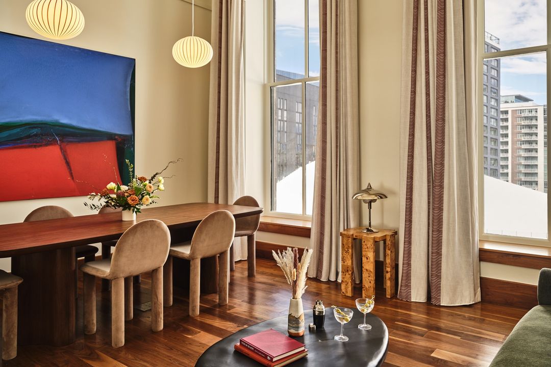 Living room interior with dining table, chairs, abstract art, coffee table with books, and cocktail glasses in a contemporary setting.