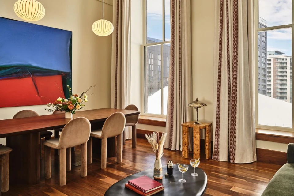 Living room interior with dining table, chairs, abstract art, coffee table with books, and cocktail glasses in a contemporary setting.
