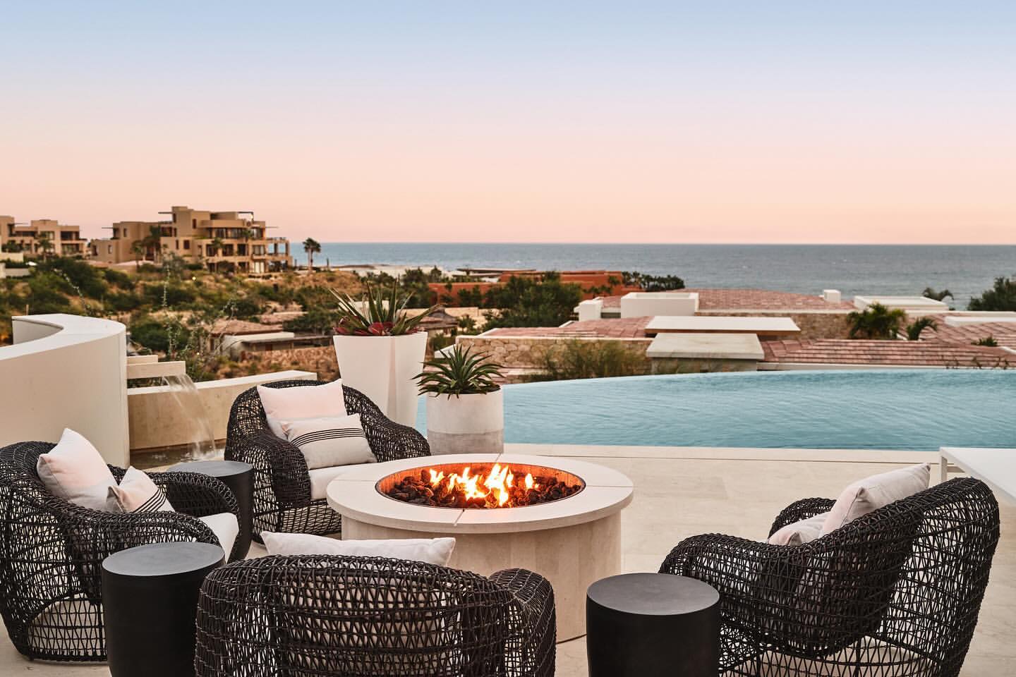 Outdoor patio with wicker chairs, side tables, and a fire pit, offering ocean views and villas in the background at dusk. Tranquil luxury.