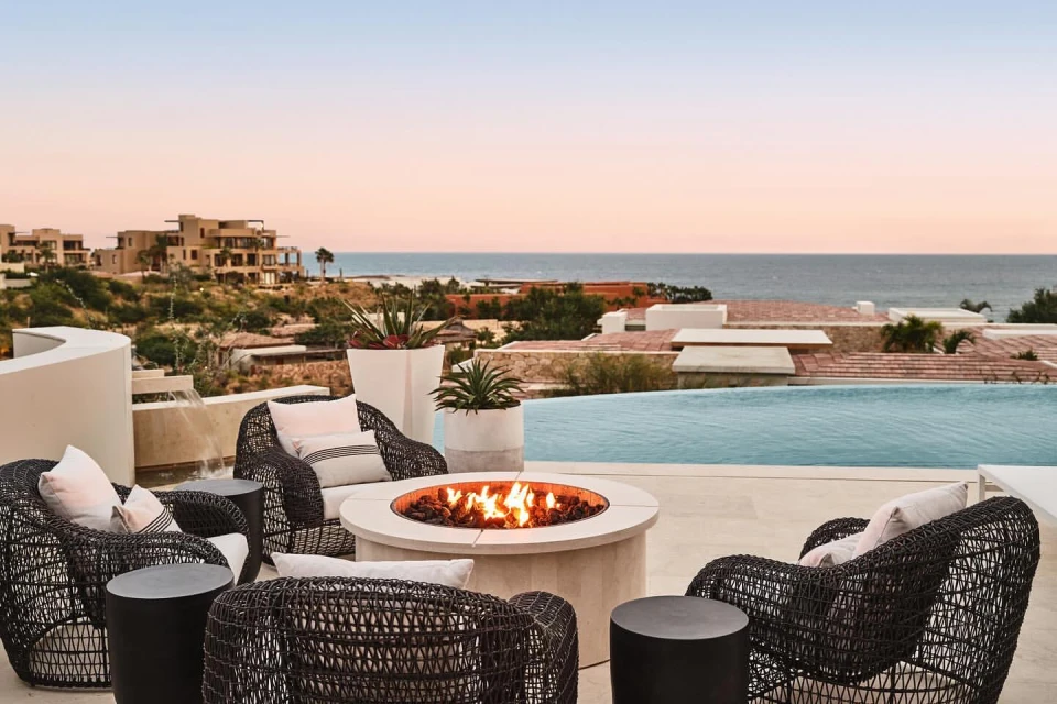 Outdoor patio with wicker chairs, side tables, and a fire pit, offering ocean views and villas in the background at dusk. Tranquil luxury.