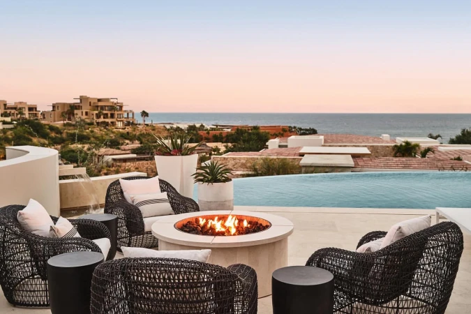 Outdoor patio with wicker chairs, side tables, and a fire pit, offering ocean views and villas in the background at dusk. Tranquil luxury.