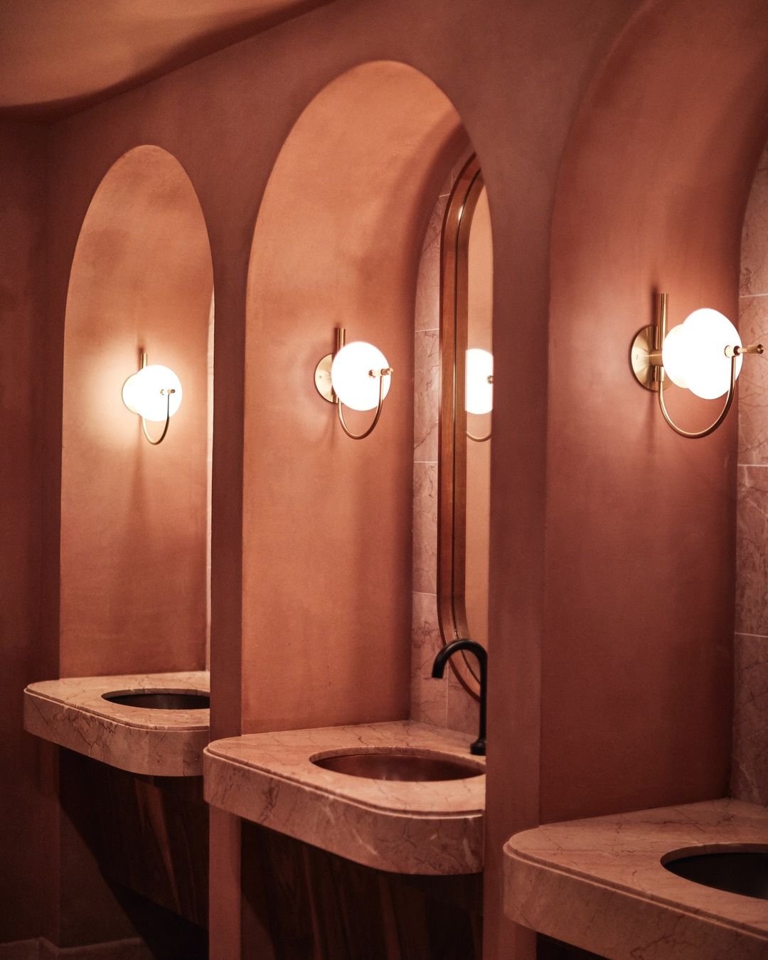 Three arched alcoves featuring marble sinks, globe sconces, and salmon-colored plaster walls create a modern, symmetrical bathroom interior design.