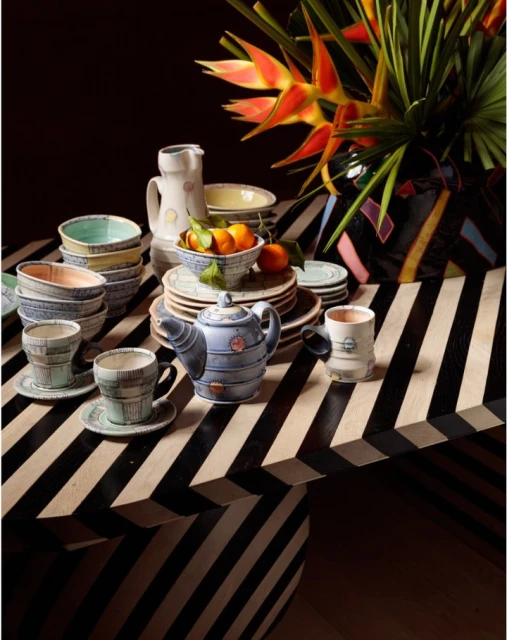 Still life on a table featuring a ceramic teapot, cups, bowls, plates, a vase with bird of paradise flowers, and tangerines.