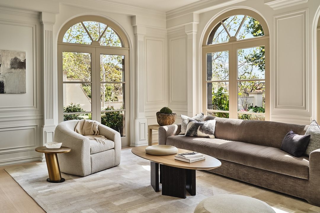 Living room interior with a brown sofa, patterned rug, modern furniture, and natural light from large arched windows overlooking greenery.
