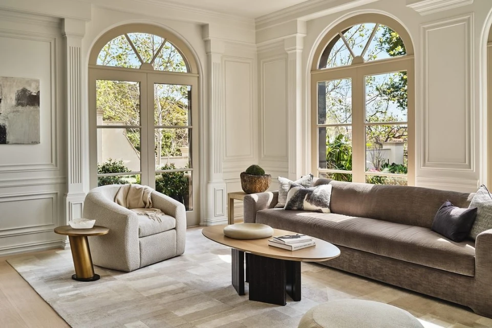 Living room interior with a brown sofa, patterned rug, modern furniture, and natural light from large arched windows overlooking greenery.
