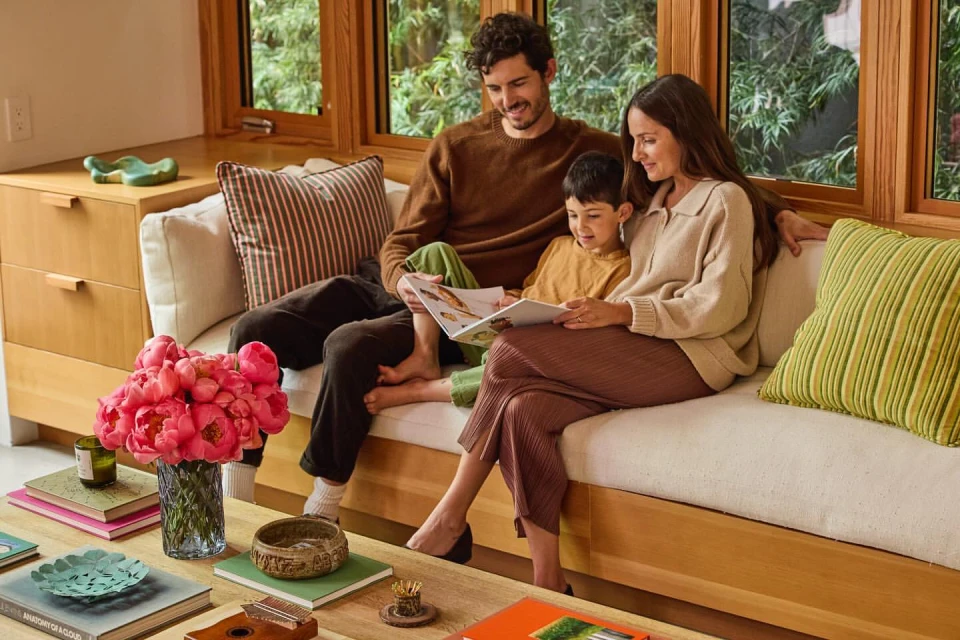 Family of three reading a book together on a cream couch in a brightly lit living room with wooden accents and a coffee table.