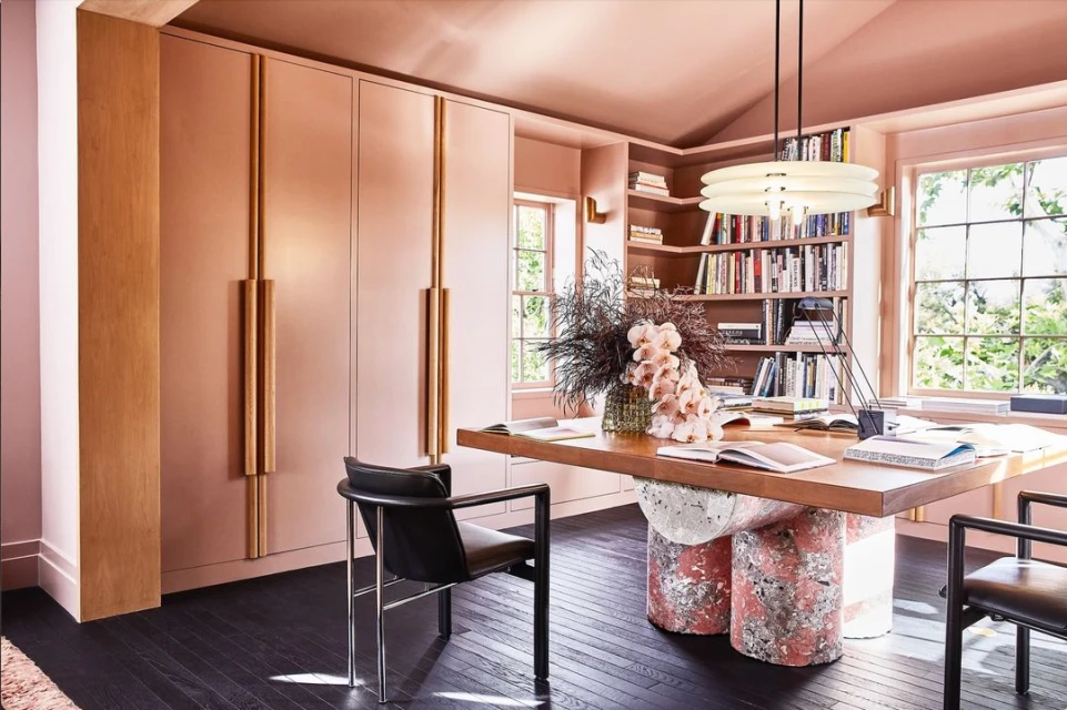 Contemporary home office interior featuring a wood desk with stone legs, upholstered chairs, and bespoke storage units in a pink-toned room.