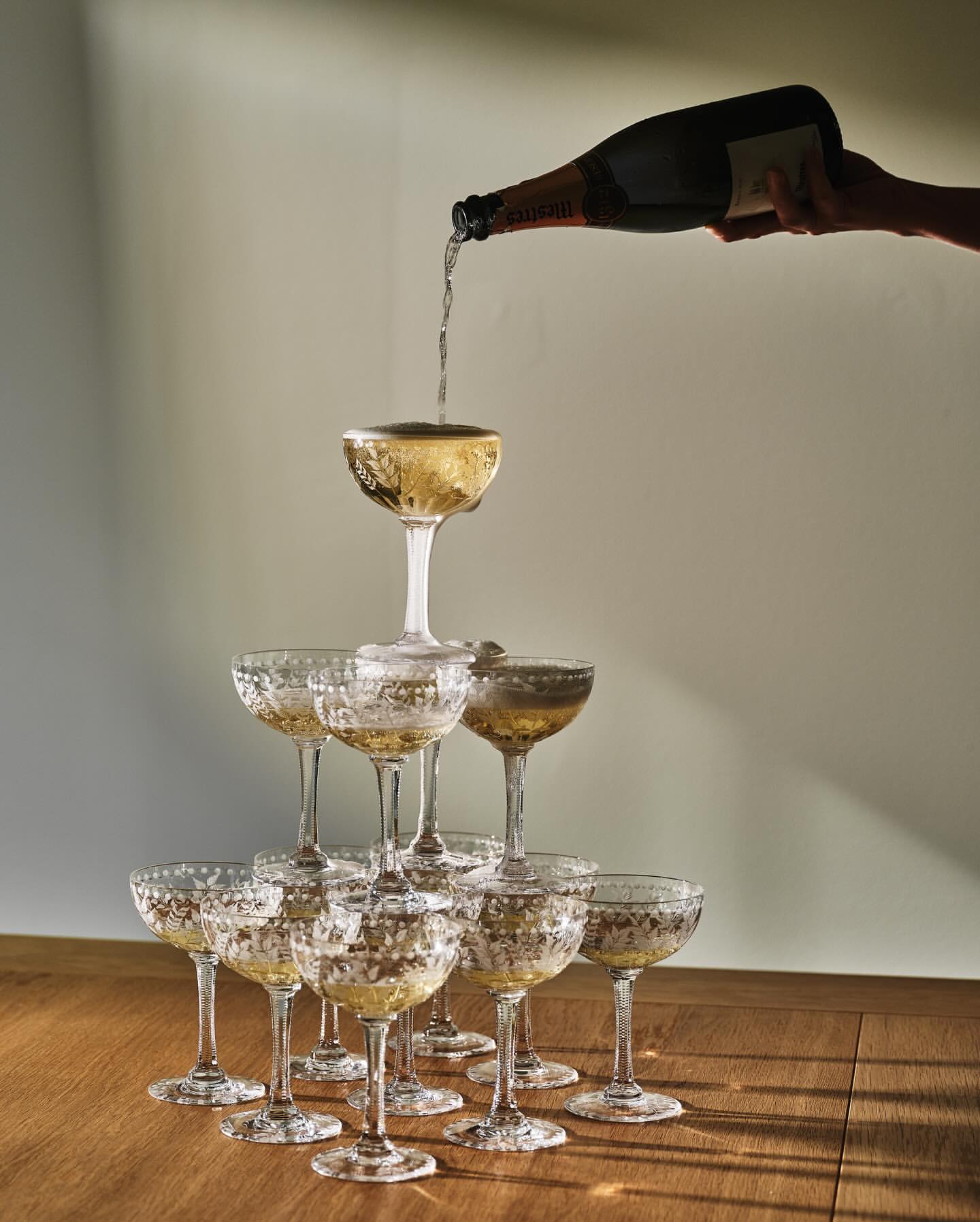 Tower of crystal champagne coupe glasses being filled with champagne from a bottle in a warm, inviting, neutral toned environment.