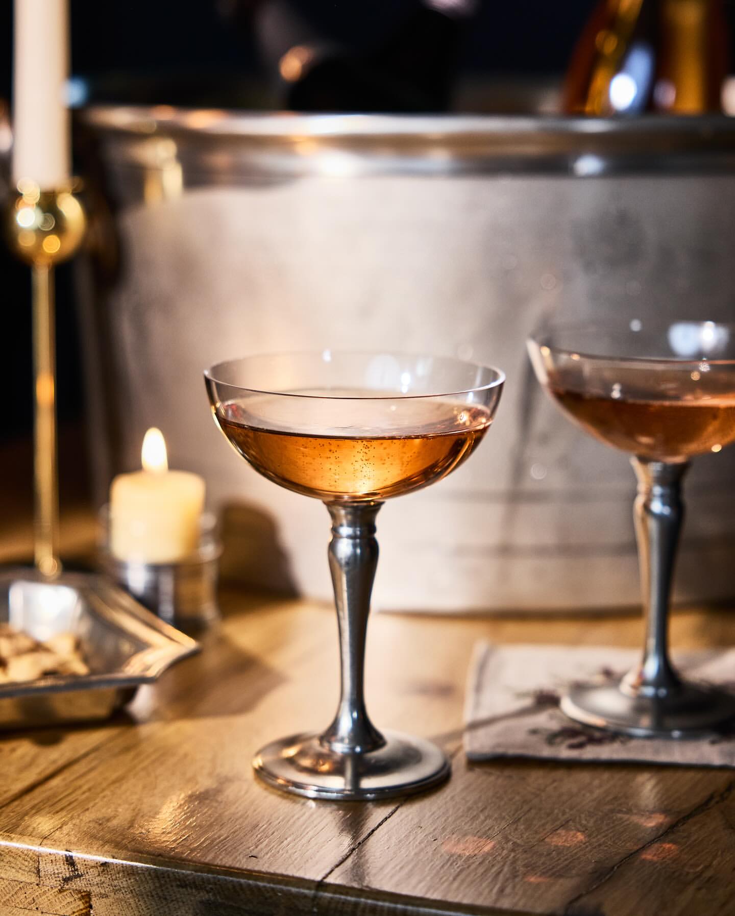 Two elegant champagne coupe glasses filled with a beverage, set on a wooden table with candles and a silver ice bucket in background.