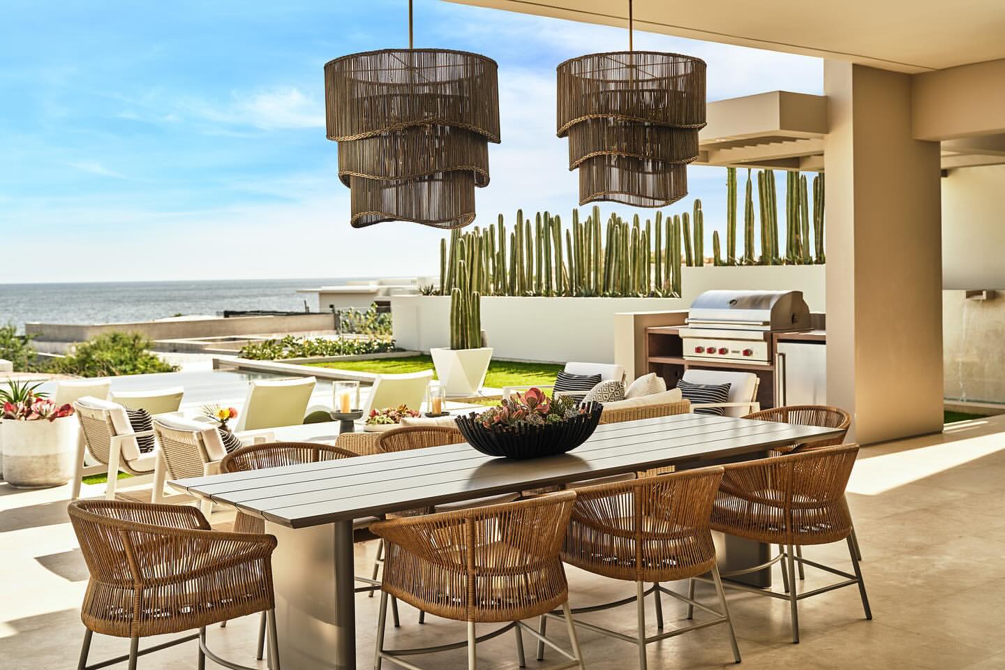 Outdoor patio with dining table and chairs, pendant lights, and desert landscaping near the ocean, creating an elegant and inviting space.