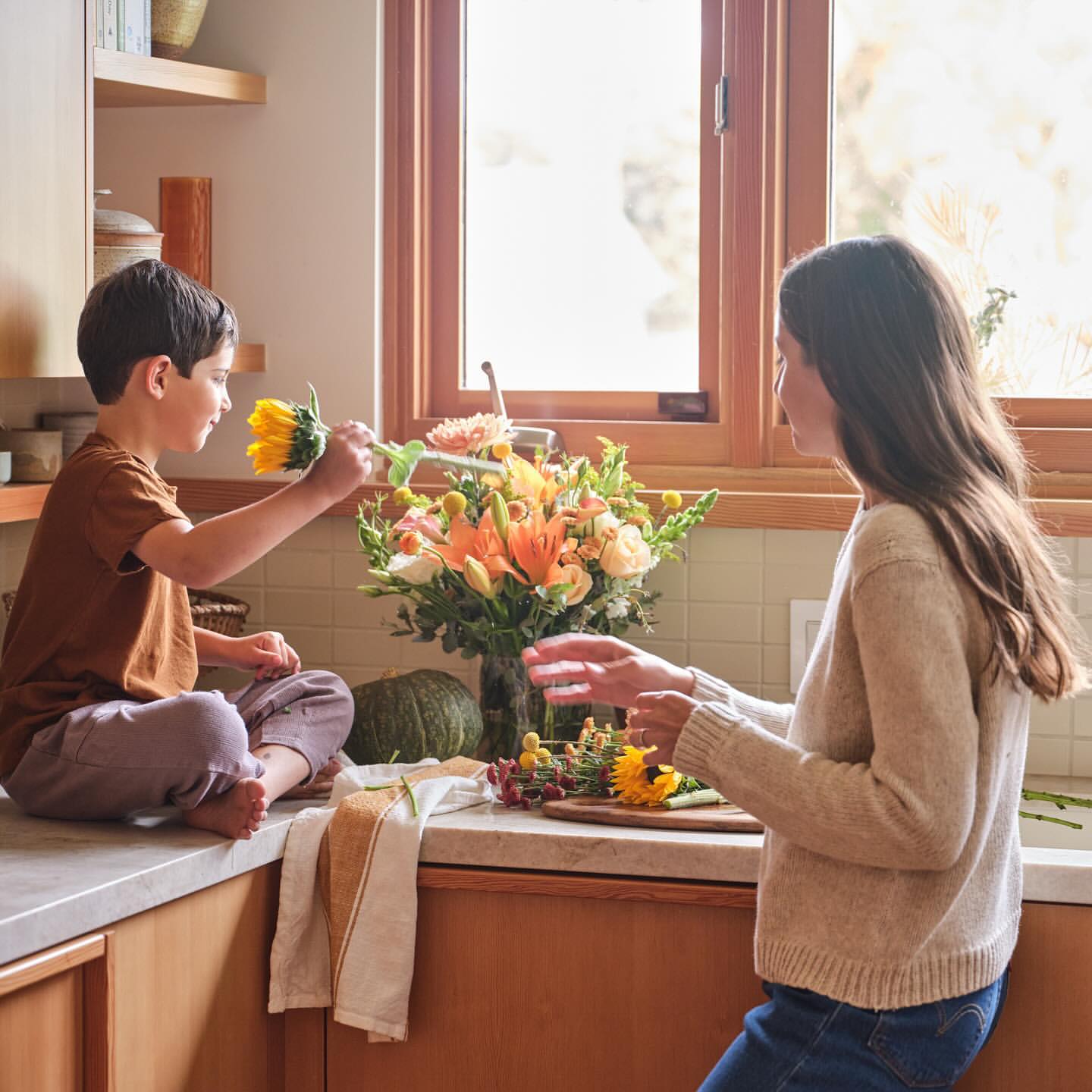 Child hands yellow sunflower to woman in kitchen. Bright floral bouquet, green pumpkin, wooden cutting board on counter.