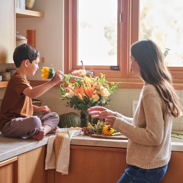 Child hands yellow sunflower to woman in kitchen. Bright floral bouquet, green pumpkin, wooden cutting board on counter.