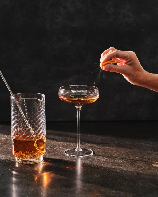 Cocktail preparation featuring a stemmed glass with a drink and a bartender's hand squeezing citrus over a dark stone surface.