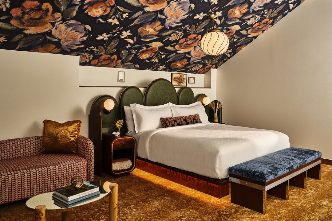 Interior of a hotel bedroom features a queen-size bed, floral-patterned ceiling, loveseat, and round accent table with books.