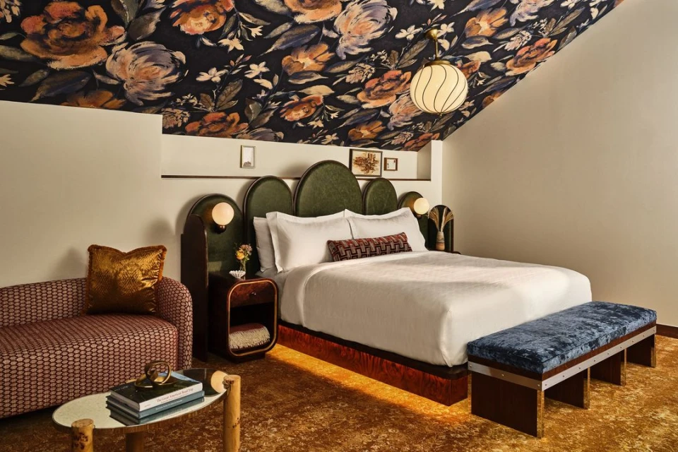 Interior of a hotel bedroom features a queen-size bed, floral-patterned ceiling, loveseat, and round accent table with books.