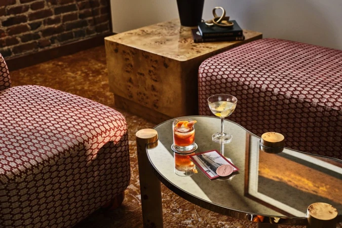 Two cocktails are on a table next to a red patterned chair, a red patterned ottoman, and a wood-grained side table in an indoor setting.