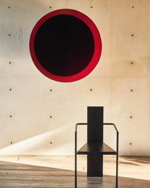 Minimalist interior with black chair against a concrete wall featuring a large red and black circle art piece above it, neutral tones.