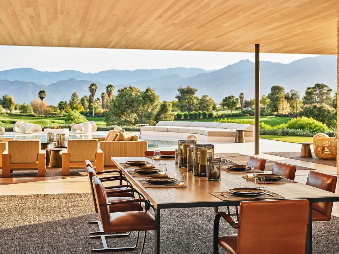 Outdoor dining area with long table set for a meal, overlooking a lush green lawn and distant mountains on a clear day.