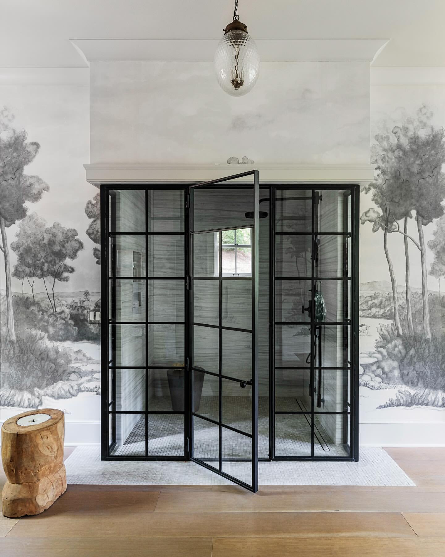 Interior bathroom features a black grid shower with glass door, light wood floors, tree stump stool, and a landscape mural wallpaper.