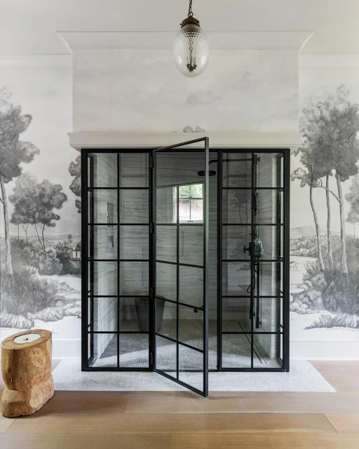 Interior bathroom features a black grid shower with glass door, light wood floors, tree stump stool, and a landscape mural wallpaper.