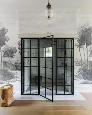 Interior bathroom features a black grid shower with glass door, light wood floors, tree stump stool, and a landscape mural wallpaper.