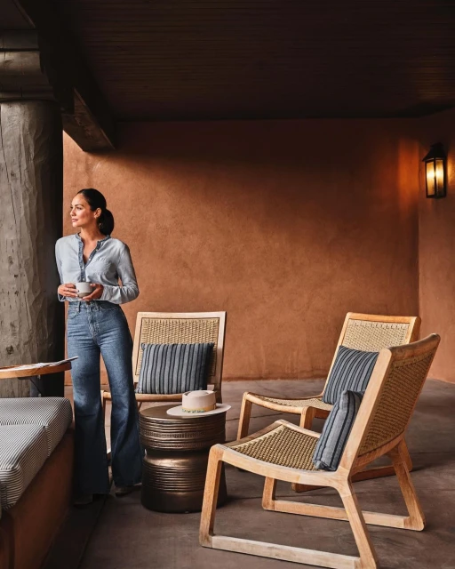 Indoor lounge area with woman holding a mug, wooden chairs with cushions, side table with hat, and earth-toned walls.