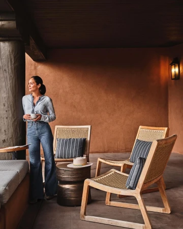 Indoor lounge area with woman holding a mug, wooden chairs with cushions, side table with hat, and earth-toned walls.