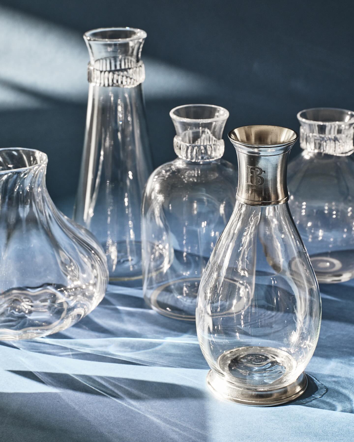 Elegant display of clear glass decanters and vases with silver accents on a blue surface, creating a sophisticated table setting.