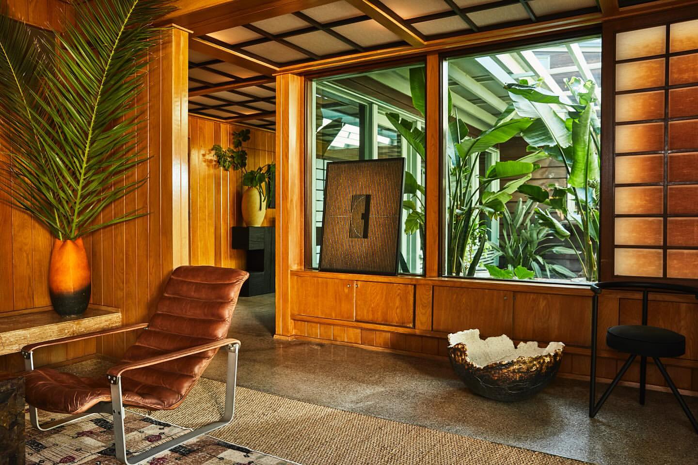 Mid-century modern living room with wood paneled walls, brown leather chair, large vase with greenery, and view through window.