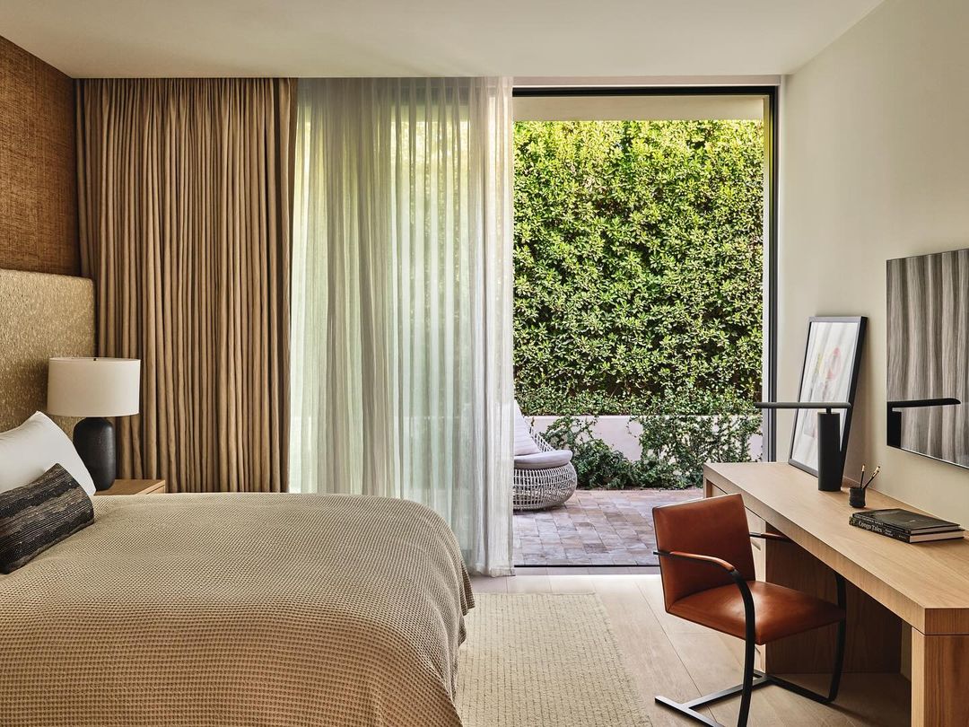 Bedroom featuring a bed, desk with chair, and view of an outdoor space with greenery, bathed in natural light and neutral colors.