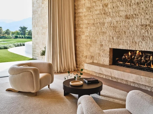 Living room interior featuring two beige boucle armchairs, a round black coffee table with decor, stone fireplace, and a view to the outside.