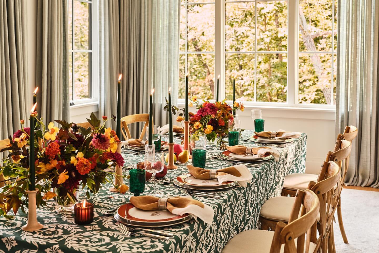 Formal dining table set with green patterned tablecloth, floral arrangements, candles, plates, silverware, and chairs.