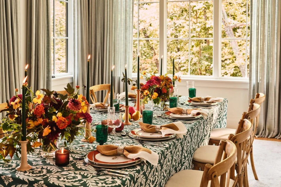 Formal dining table set with green patterned tablecloth, floral arrangements, candles, plates, silverware, and chairs.