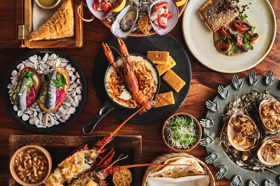 Overhead shot of a wooden table laden with a variety of seafood dishes including oysters, grilled fish, lobster, and various sides.