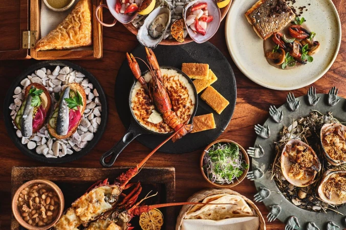 Overhead shot of a wooden table laden with a variety of seafood dishes including oysters, grilled fish, lobster, and various sides.
