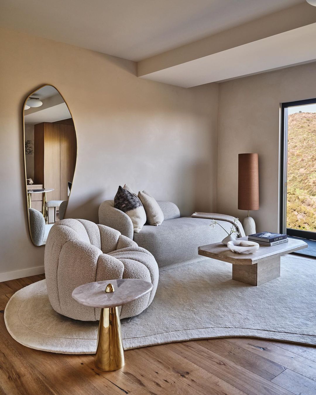 Interior design featuring a curved sofa and armchairs on a light rug, accented by a gold side table, floor lamp, and view from the window.