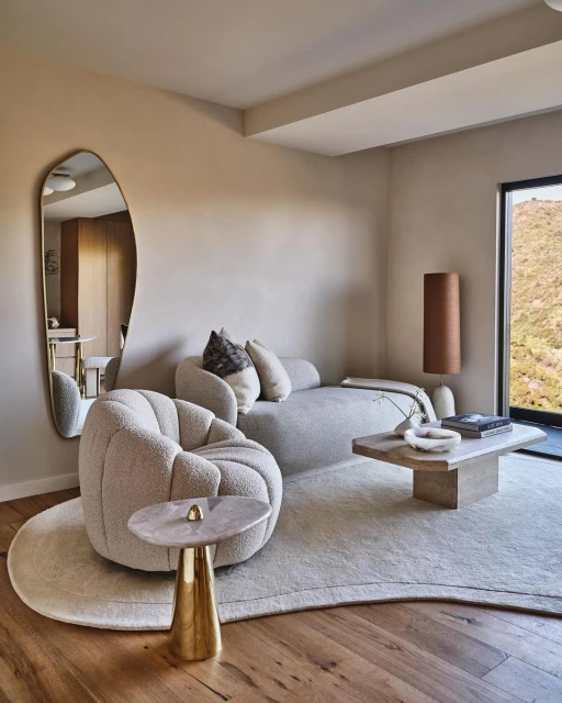 Interior design featuring a curved sofa and armchairs on a light rug, accented by a gold side table, floor lamp, and view from the window.