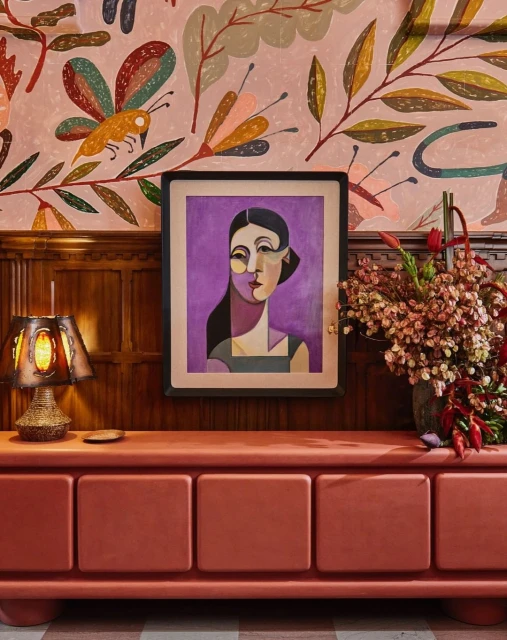 Eclectic interior with floral wallpaper, abstract portrait art, lamp, and salmon-colored credenza against wood paneling. Interior design.