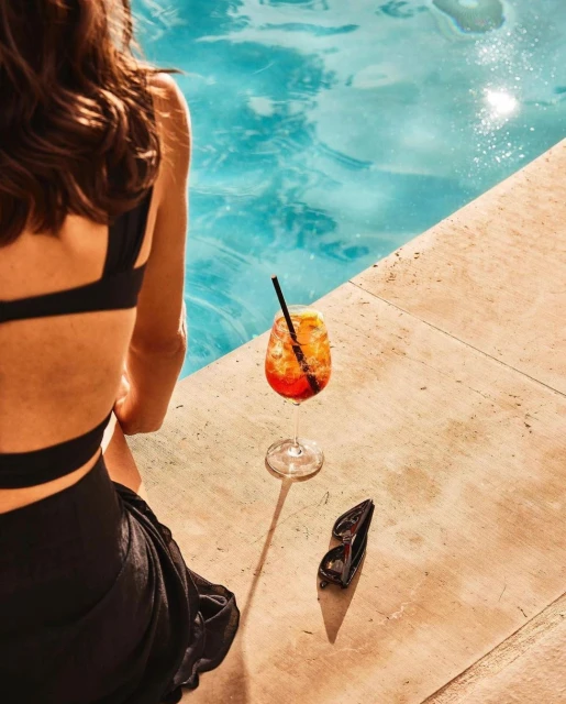 Woman sits poolside near cocktail and sunglasses, showcasing a relaxed summer scene with refreshing drink and accessories in outdoor setting.