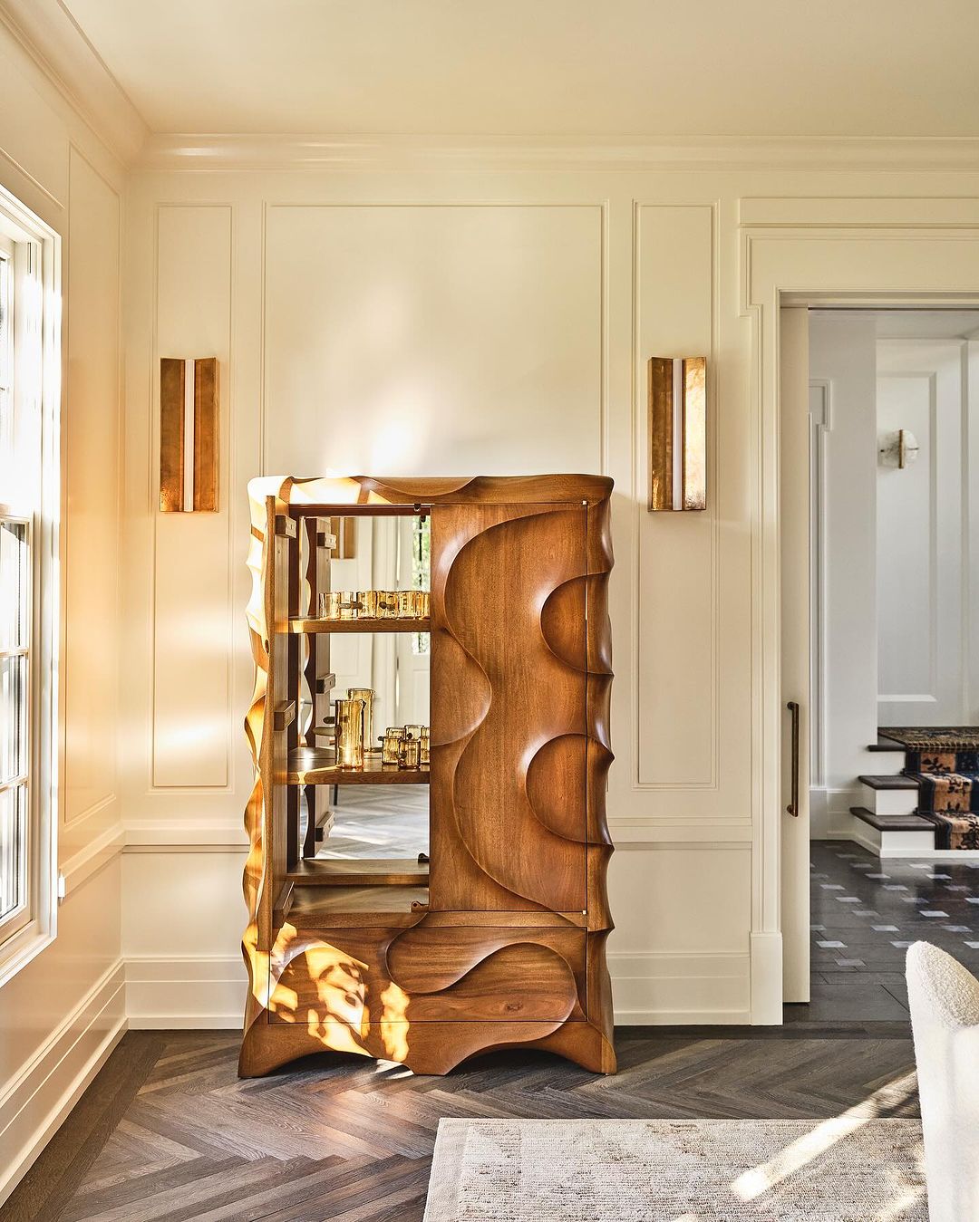 Modern wood and mirrored bar cabinet featuring curved accents and multiple shelves, illuminated by sconce lighting in a cream-colored room.