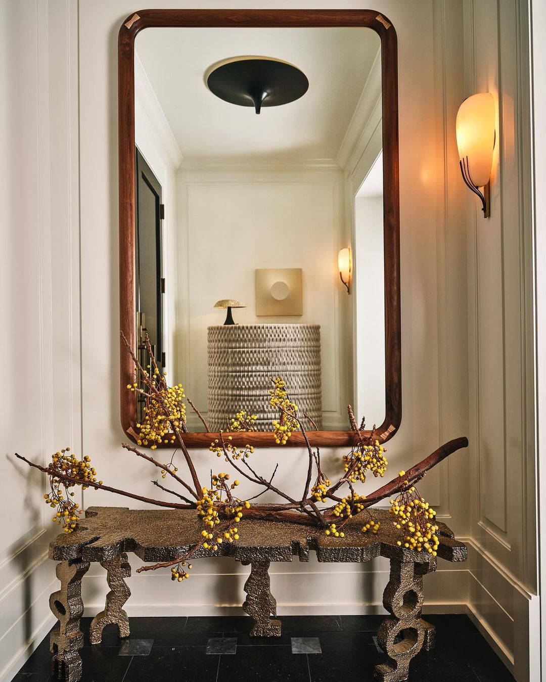 Interior hallway with a wooden framed mirror above a console table decorated with yellow berries in front of a white wall.