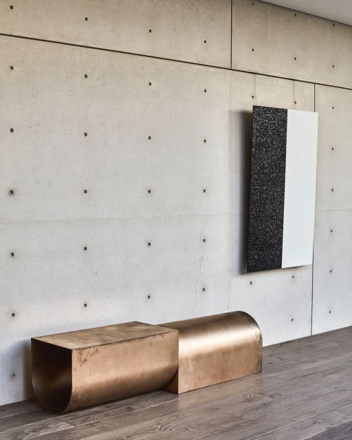 Contemporary interior with bronze metal bench, abstract art hanging on a concrete wall, and grey hardwood flooring in a modern architectural space.