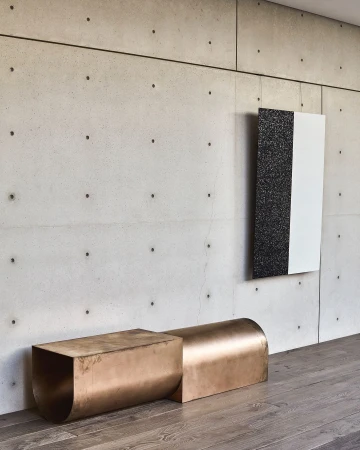Contemporary interior with bronze metal bench, abstract art hanging on a concrete wall, and grey hardwood flooring in a modern architectural space.