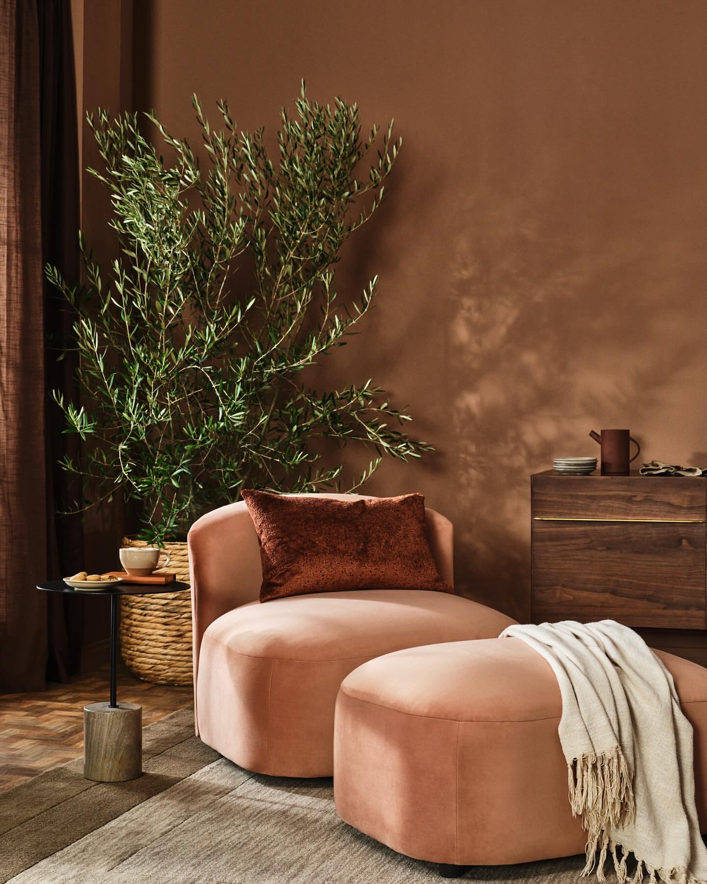 Interior photograph featuring a soft pink chair and ottoman, a tall green plant, a round side table, and a wooden cabinet.