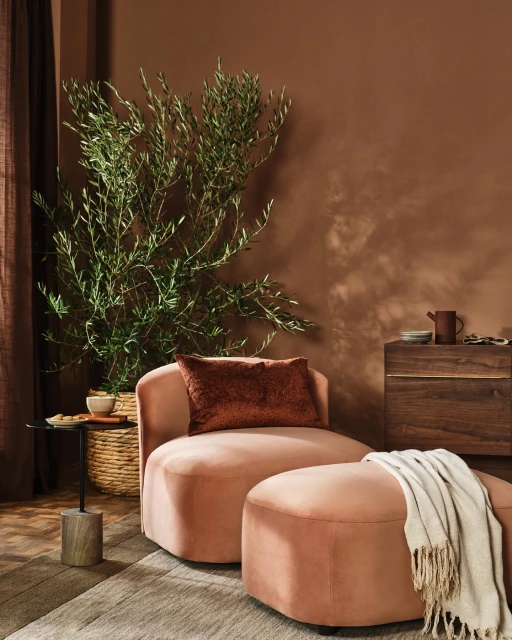 Interior photograph featuring a soft pink chair and ottoman, a tall green plant, a round side table, and a wooden cabinet.